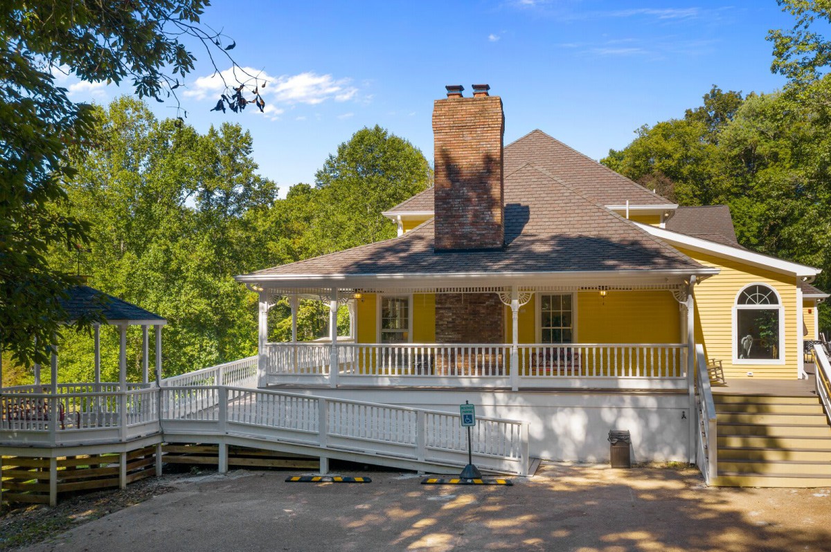 A yellow house with a spacious porch and a chimney, surrounded by trees on a sunny day.