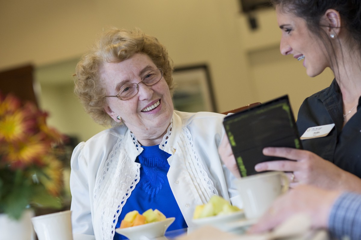 An elderly woman smiling while looking at a tablet during a conversation.