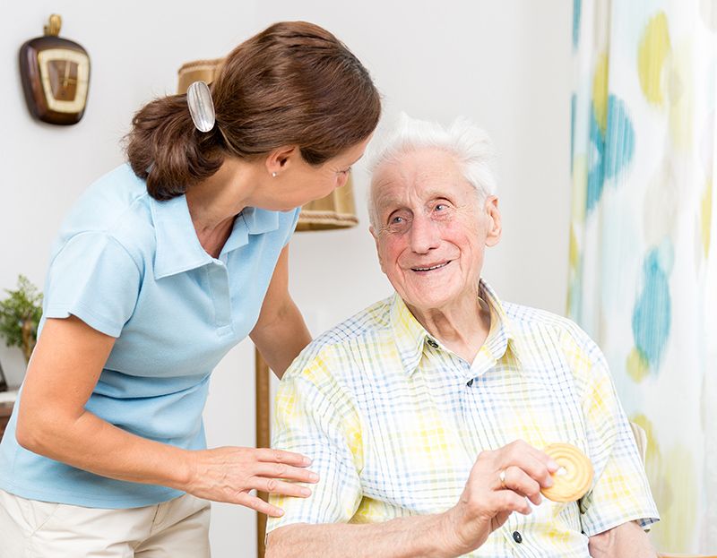 A caregiver smiling and interacting with an elderly man in a home setting.