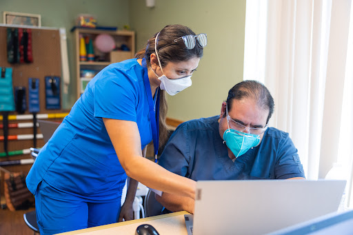 A healthcare professional assists a patient with using a laptop in a medical setting.