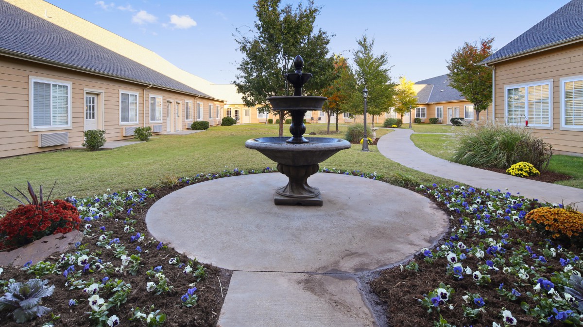 A serene courtyard with a central fountain, surrounded by landscaped gardens and winding pathways, flanked by buildings.