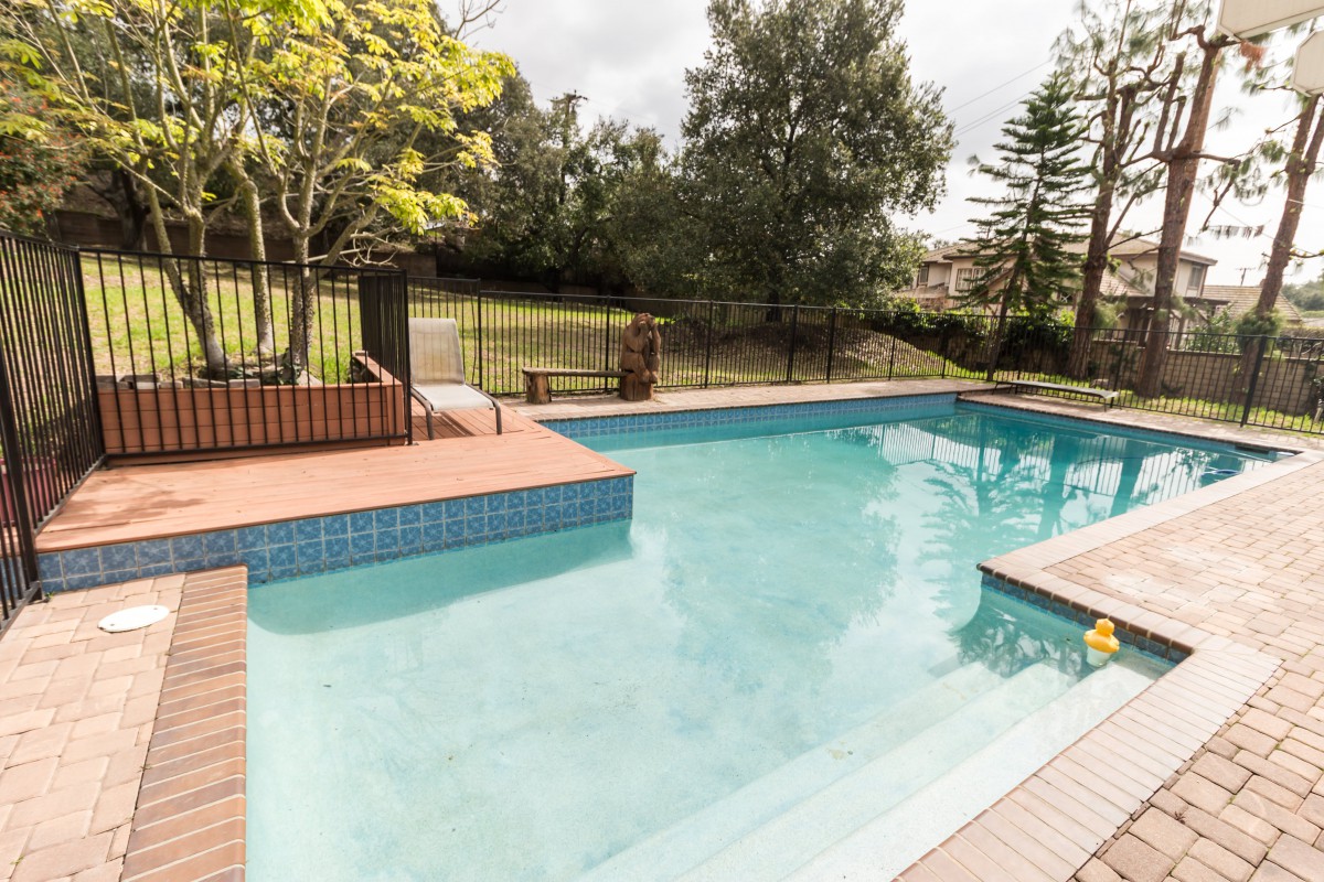 A serene outdoor swimming pool surrounded by trees and a safety fence on a sunny day.
