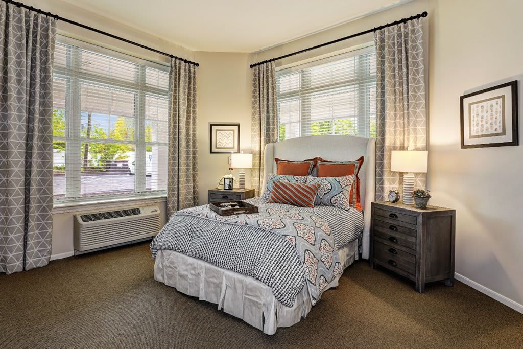 A cozy and elegantly decorated bedroom featuring a neatly made bed, large windows with curtains, bedside tables with lamps, and decorative pillows.