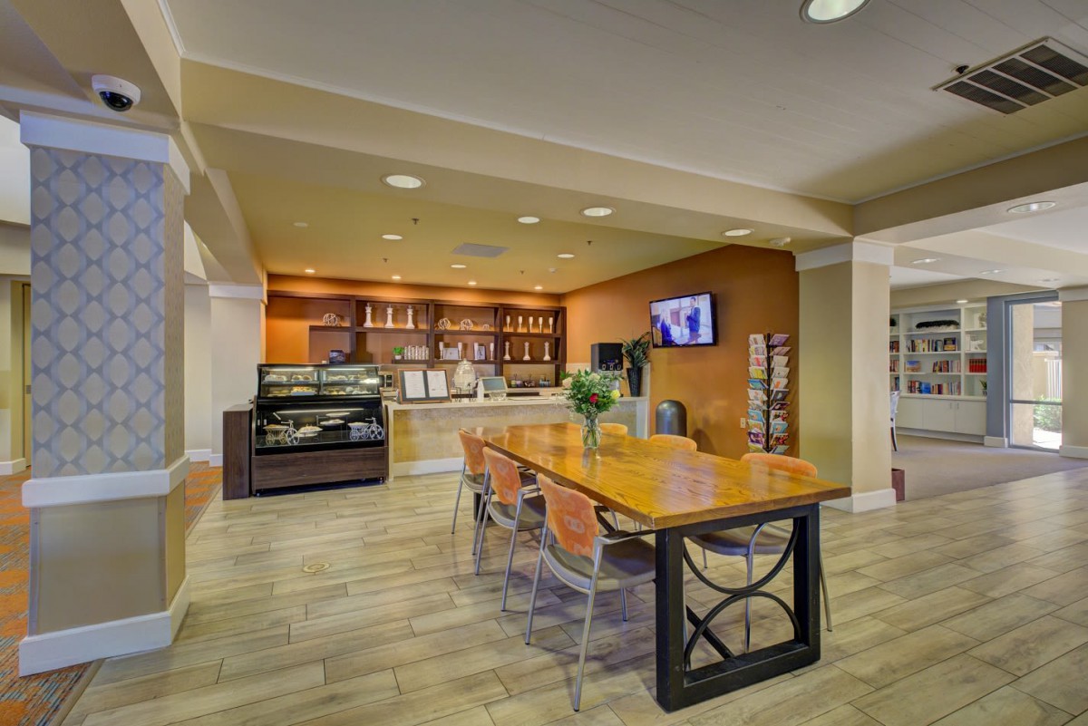 A modern cafe with a seating area featuring a long table, chairs, and a television on the wall.