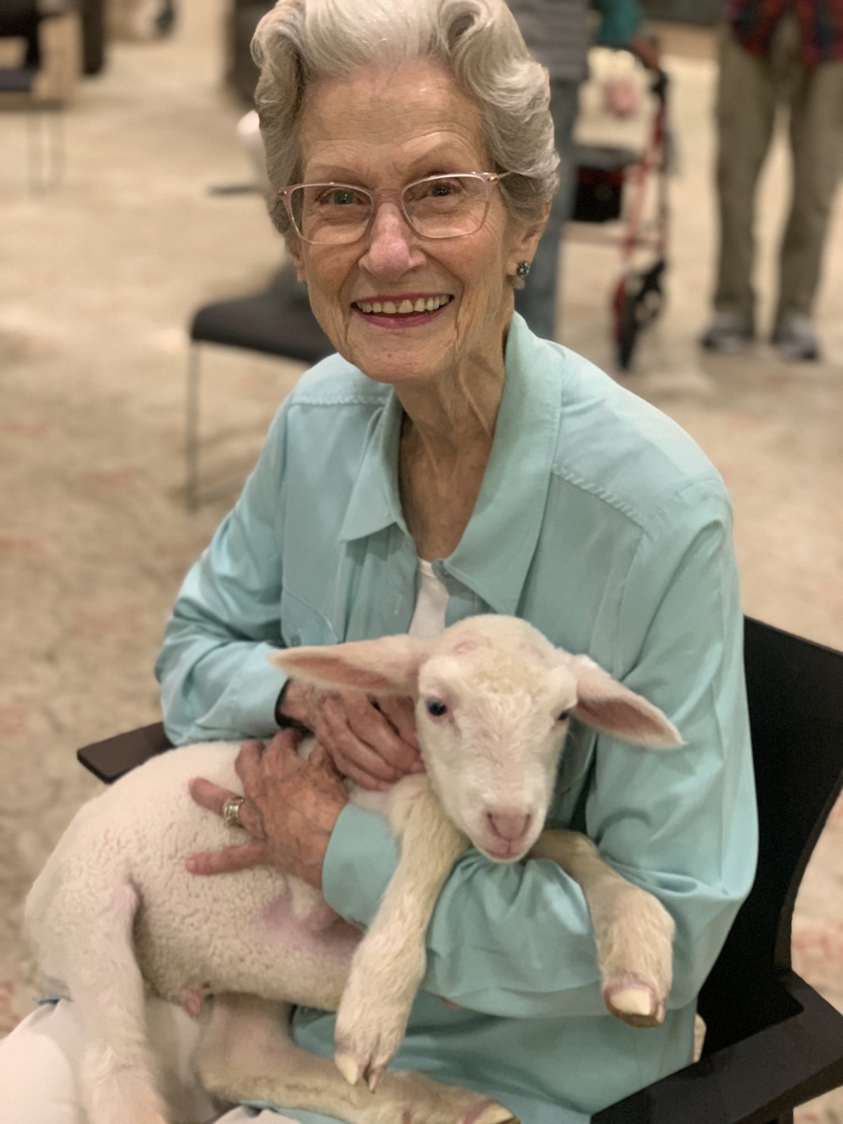 An elderly woman smiling while holding a lamb indoors.