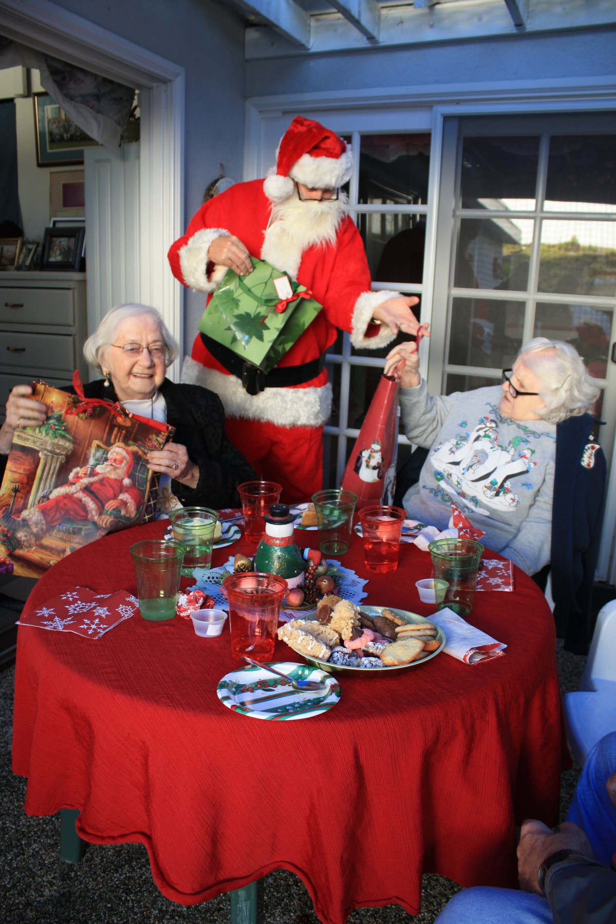 Two elderly people share a joyful moment with Santa, exchanging gifts at a festive Christmas table.
