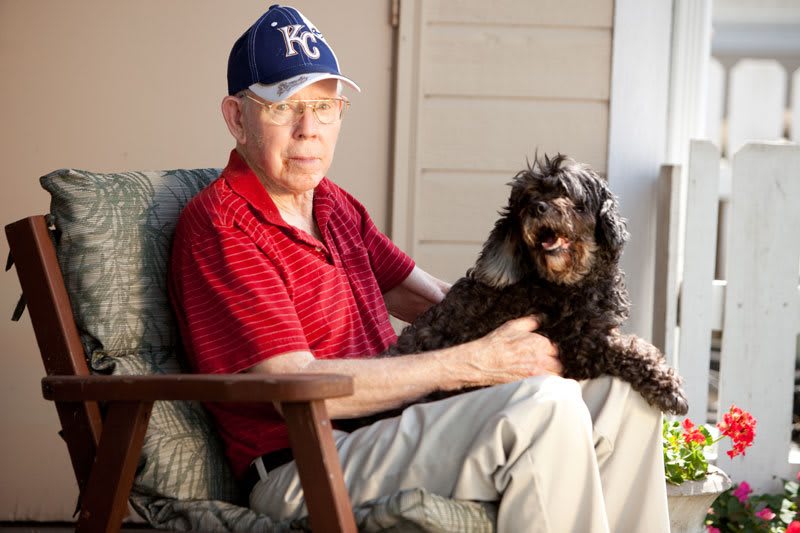 An elderly man wearing a hat sits in a chair with a small dog on his lap.