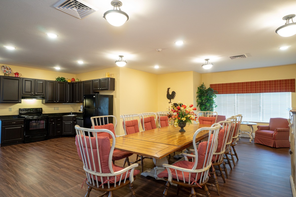A cozy dining area featuring a long wooden table surrounded by cushioned chairs, adjacent to a kitchen space with dark cabinetry.
