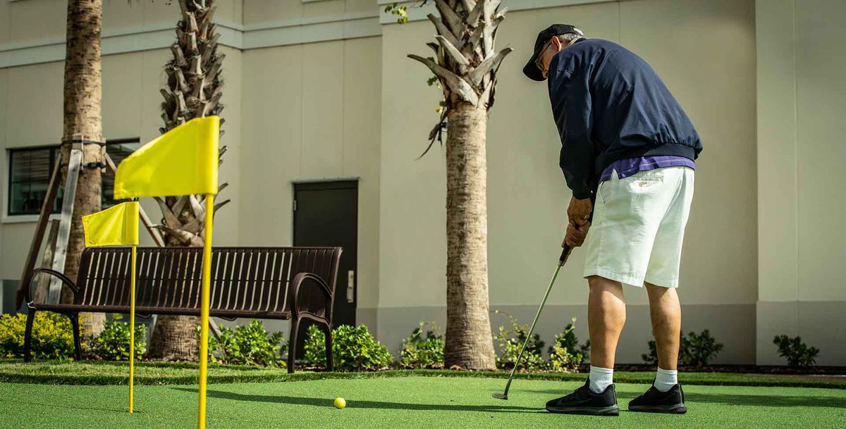 A person practicing putting on an outdoor green, near a bench and palm tree.