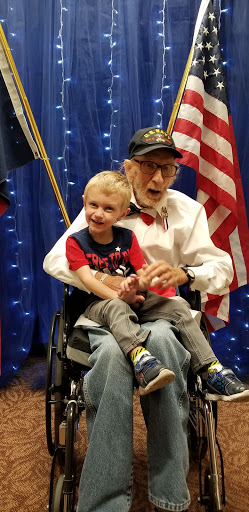 A heartwarming moment shared between an elderly man in a wheelchair and a young boy, both smiling against a backdrop of American flags.