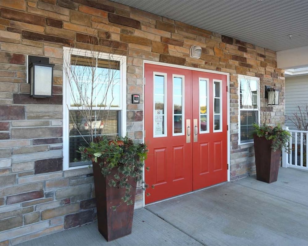 A building entrance featuring a bright red double door set in a stone wall, with potted plants and windows on either side.