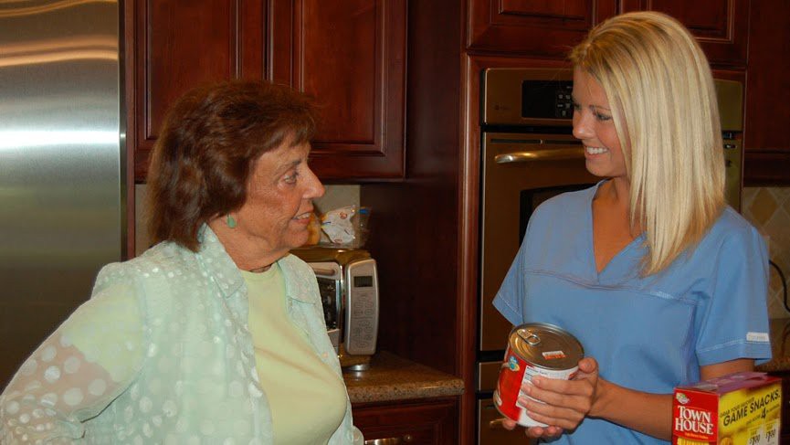 An elderly woman and a younger woman are having a conversation in a kitchen, with the younger woman holding a gift.