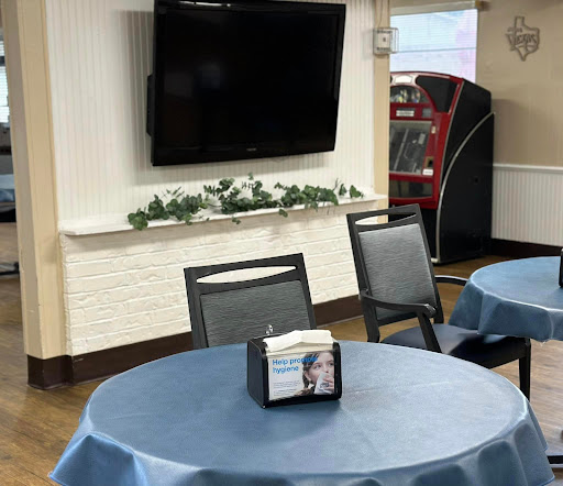 A cozy dining area featuring tables with blue tablecloths, a wall-mounted TV, and a vending machine in the background.