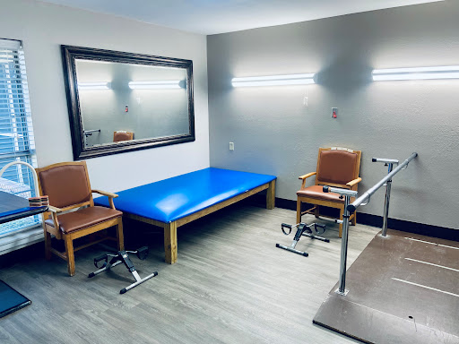 A view of a physical therapy treatment room featuring a blue table, two chairs, and various rehabilitation equipment.