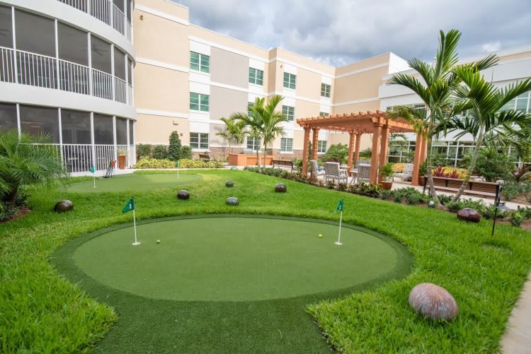 A neatly landscaped outdoor area with a small golf putting green and a pergola, adjacent to a modern building.