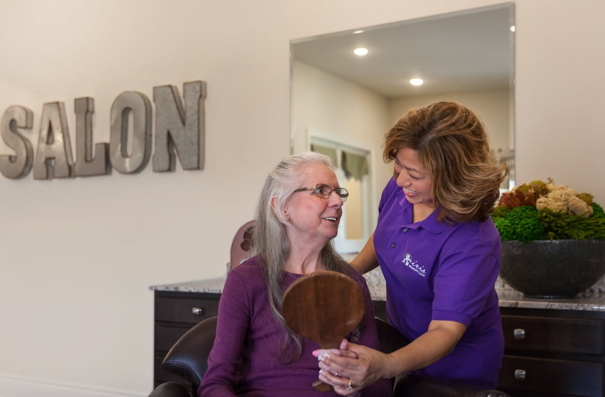 A stylist assists an elderly woman in a salon, holding a mirror to show her hairstyle.
