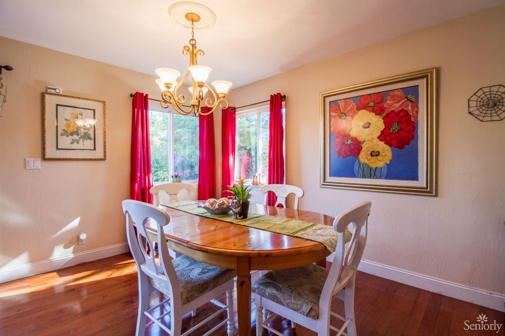A cozy dining room featuring a wooden table, chairs, chandelier lighting, vibrant red curtains, and colorful artwork.
