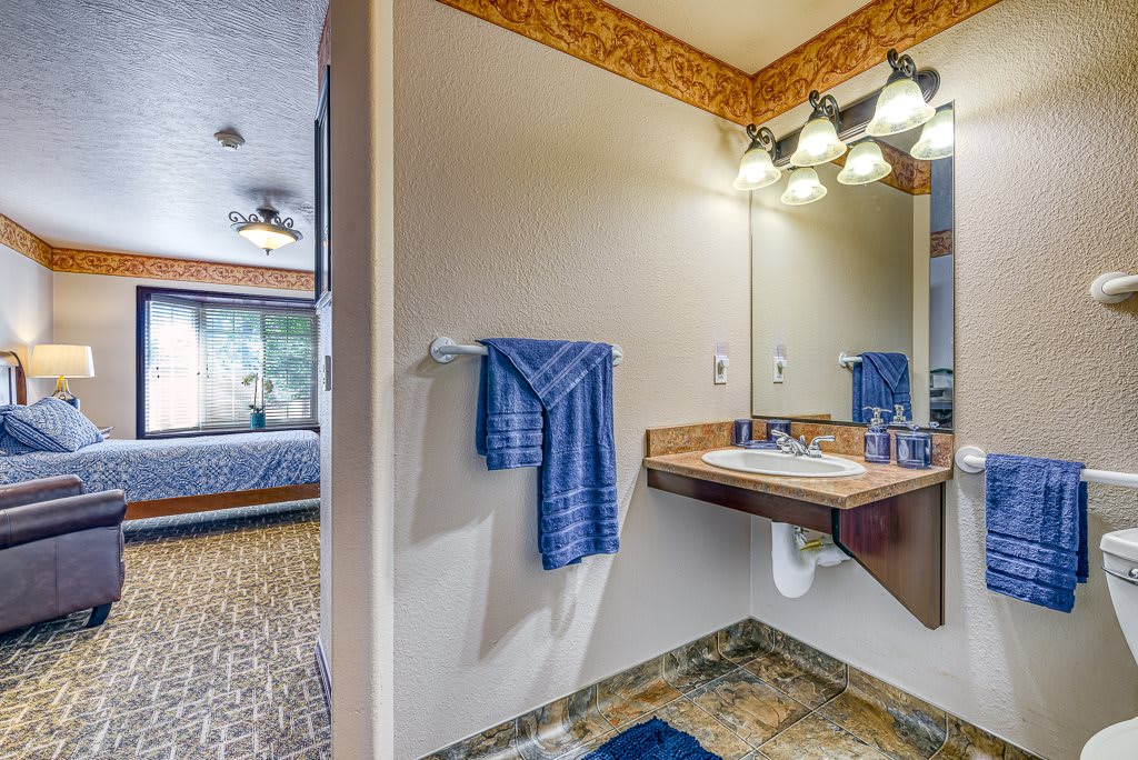 A cozy bathroom interior featuring a sink with a mirror, blue towel, decorative lighting, and a glimpse of a bedroom in the background.