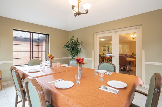 A neatly arranged dining room with an orange tablecloth, set with plates and cutlery, featuring a vase of flowers in the center.