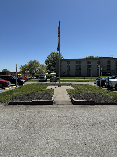 A clear day showcasing a flag in front of a parking area, flanked by neatly trimmed grass and a distant building.