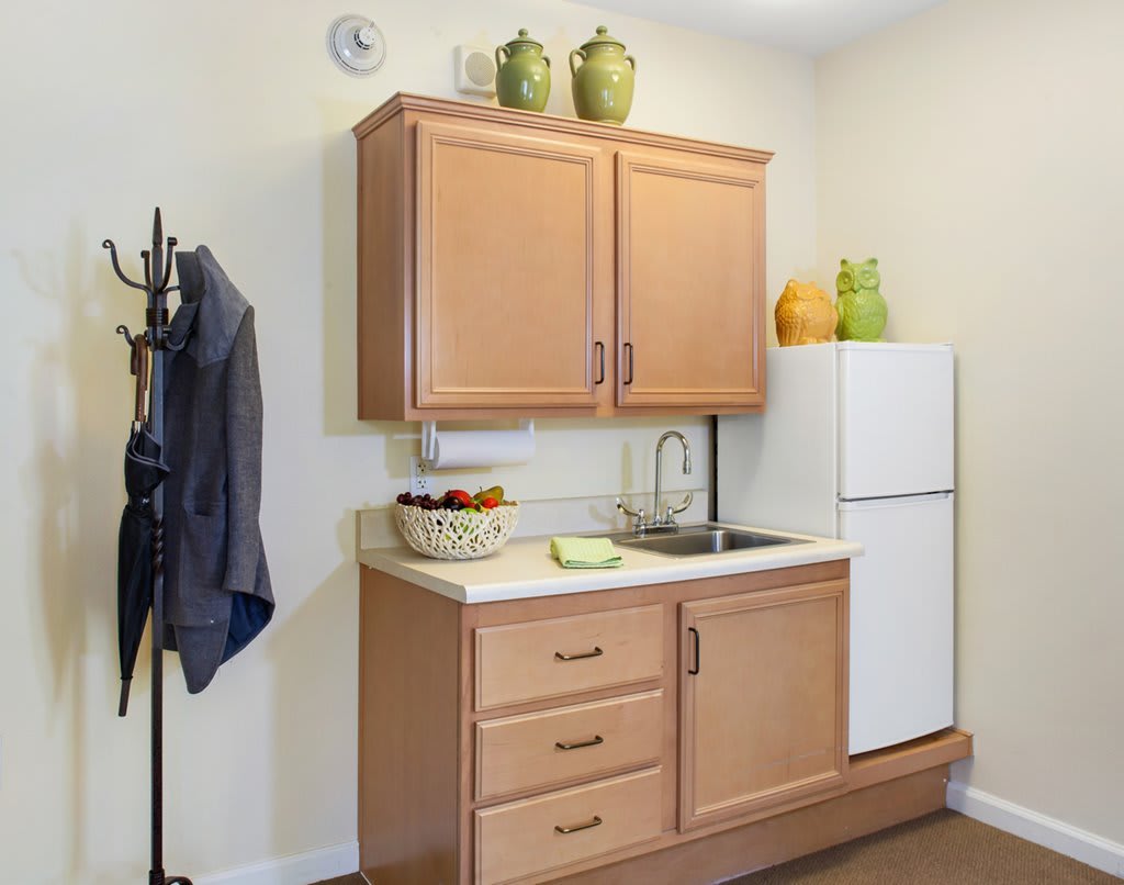 A small kitchen with wooden cabinets, a sink, and a white refrigerator. Decorative green vases are displayed above the cabinetry alongside a fruit bowl on the counter.