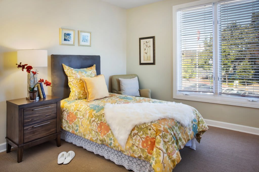 A cozy bedroom with a single bed and colorful bedding, featuring a window with blinds and a small nightstand.