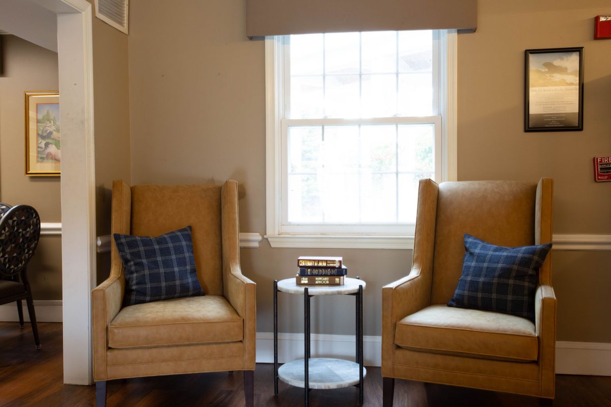 A cozy living room setup featuring two beige armchairs with plaid pillows, a small round side table topped with books, and a window letting in natural light.