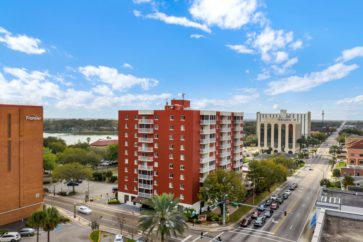 A red brick apartment building situated at a busy intersection in an urban area, with surrounding greenery and a clear blue sky.