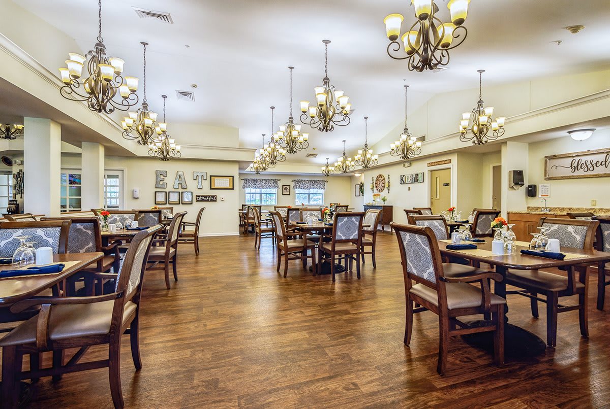 An elegant dining room with wooden floors, furnished with multiple tables and chairs, illuminated by ornate chandeliers.