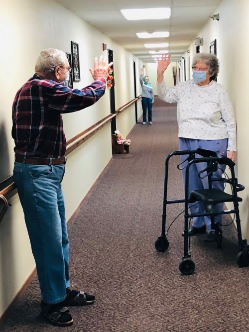 Two elderly individuals greet each other with a high five in a hallway, both wearing masks and one using a walker.