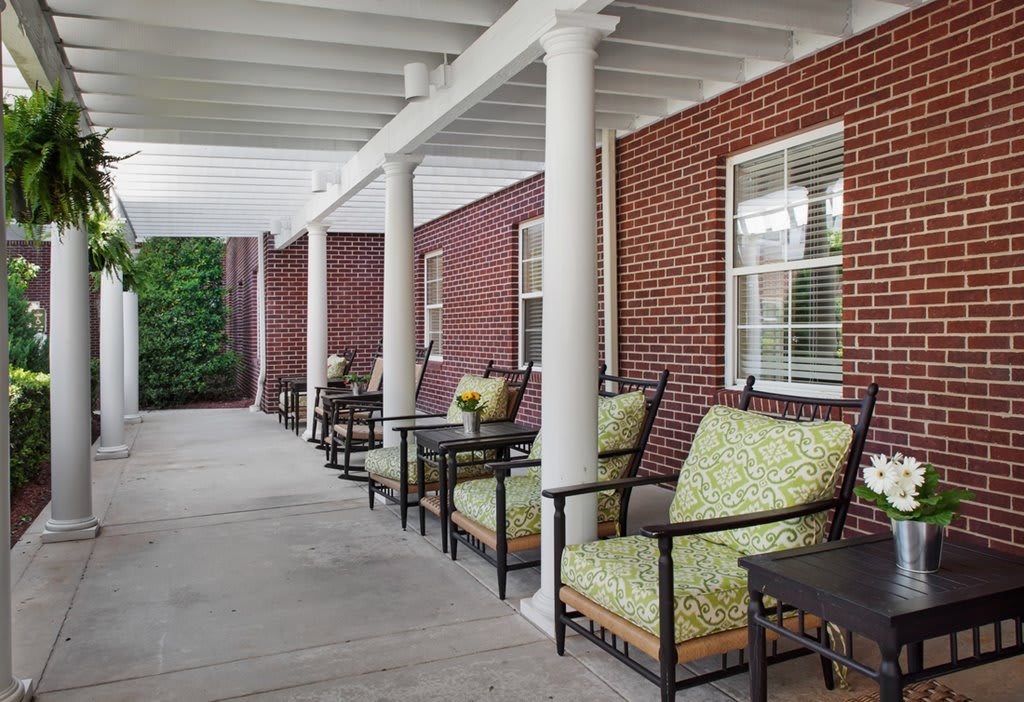 A covered porch with comfortable outdoor seating, featuring cushioned chairs and tables along a brick wall.