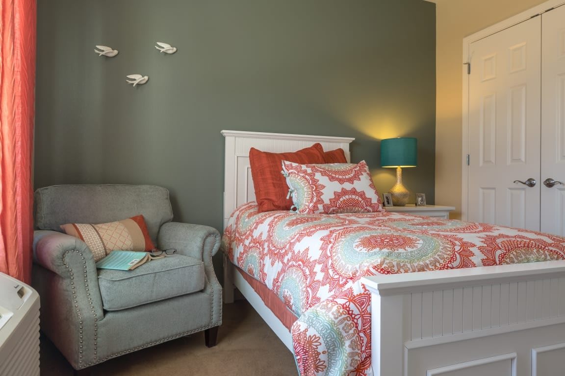A cozy bedroom with a patterned comforter on a white bed, accompanied by an armchair and lamp.