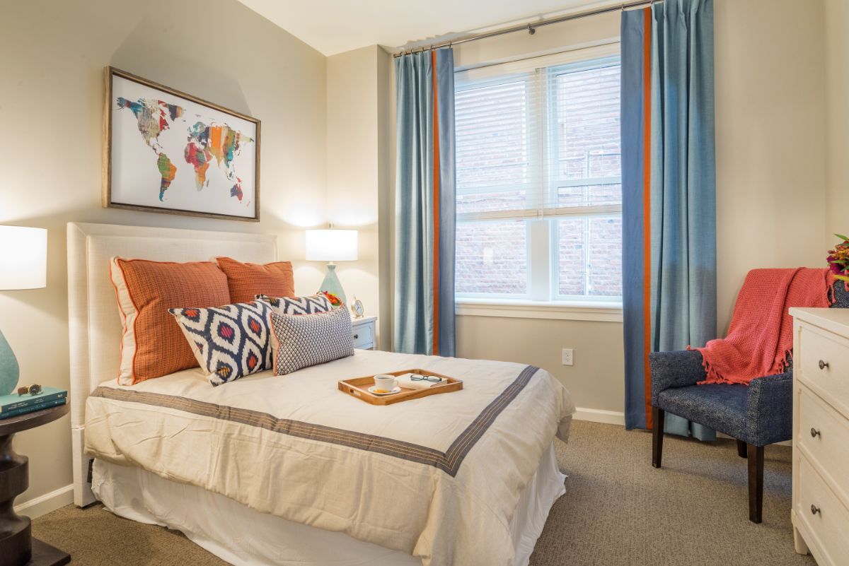 A modern and cozy bedroom with stylish decor, featuring a neatly made bed with colorful pillows, large windows with elegant curtains, and a comfortable armchair.