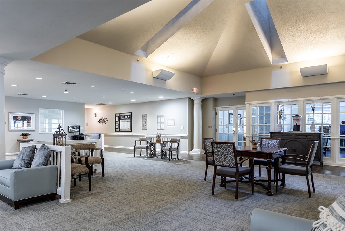 A spacious and well-lit dining area with multiple tables and chairs, featuring a carpeted floor and large windows.