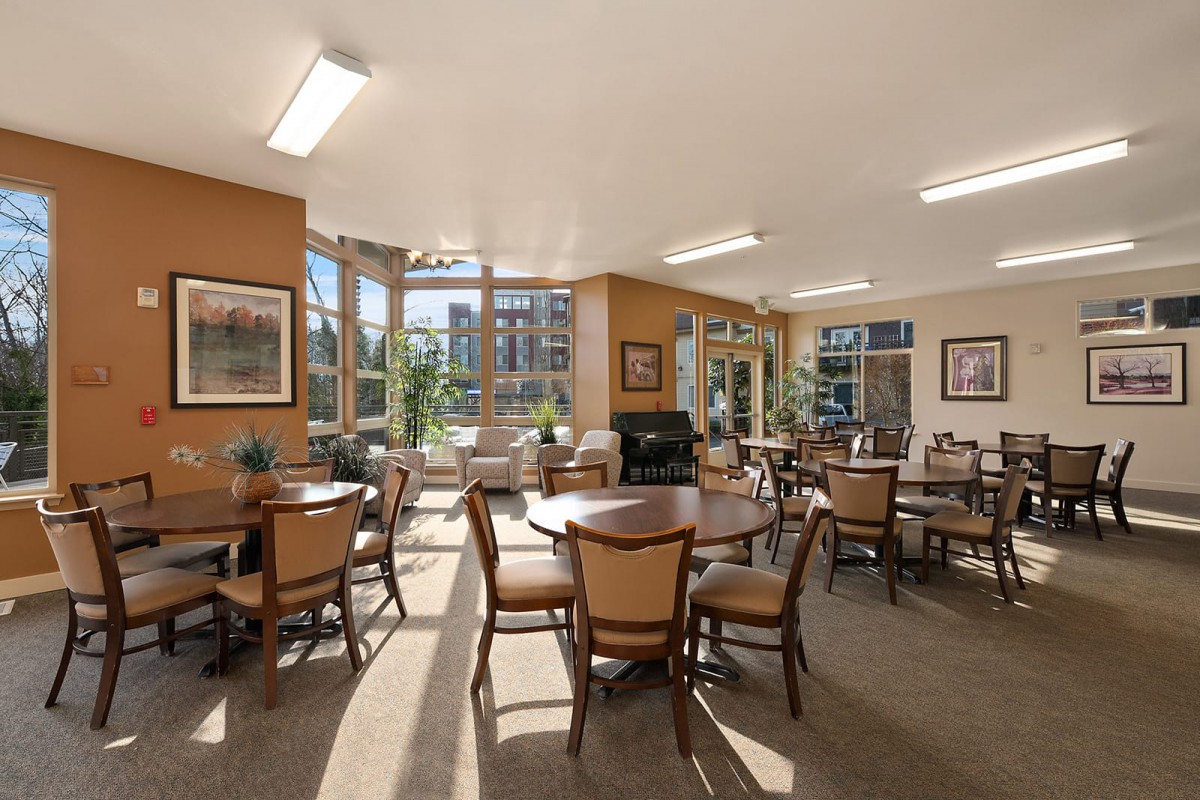 A spacious lounge area with multiple tables and chairs, sunlit through large windows.