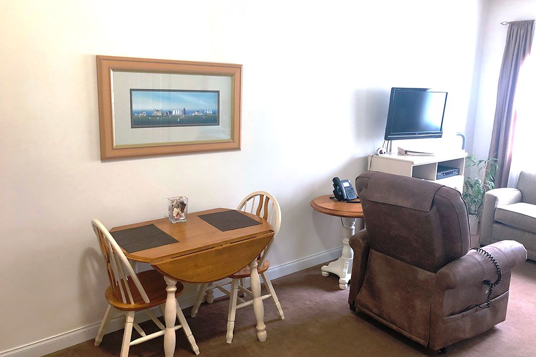 A cozy living space featuring a small dining table with chairs, a painting on the wall, and a TV setup next to an armchair.