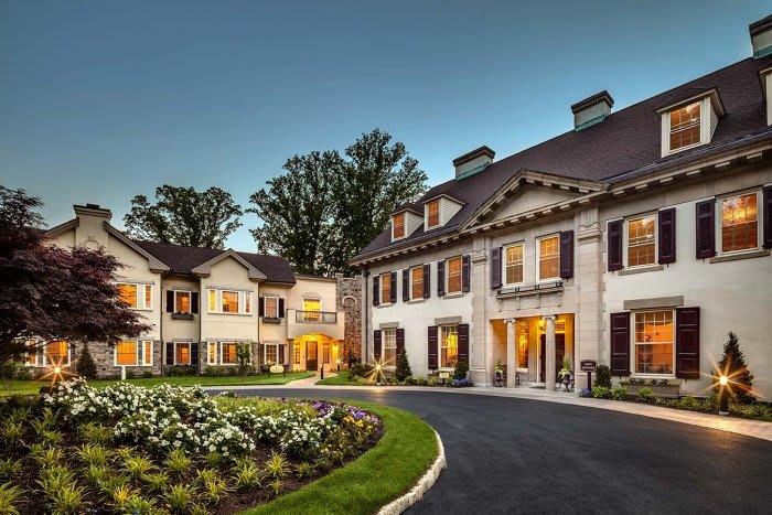 A grand luxury mansion illuminated during the evening with landscaped gardens and a circular driveway.