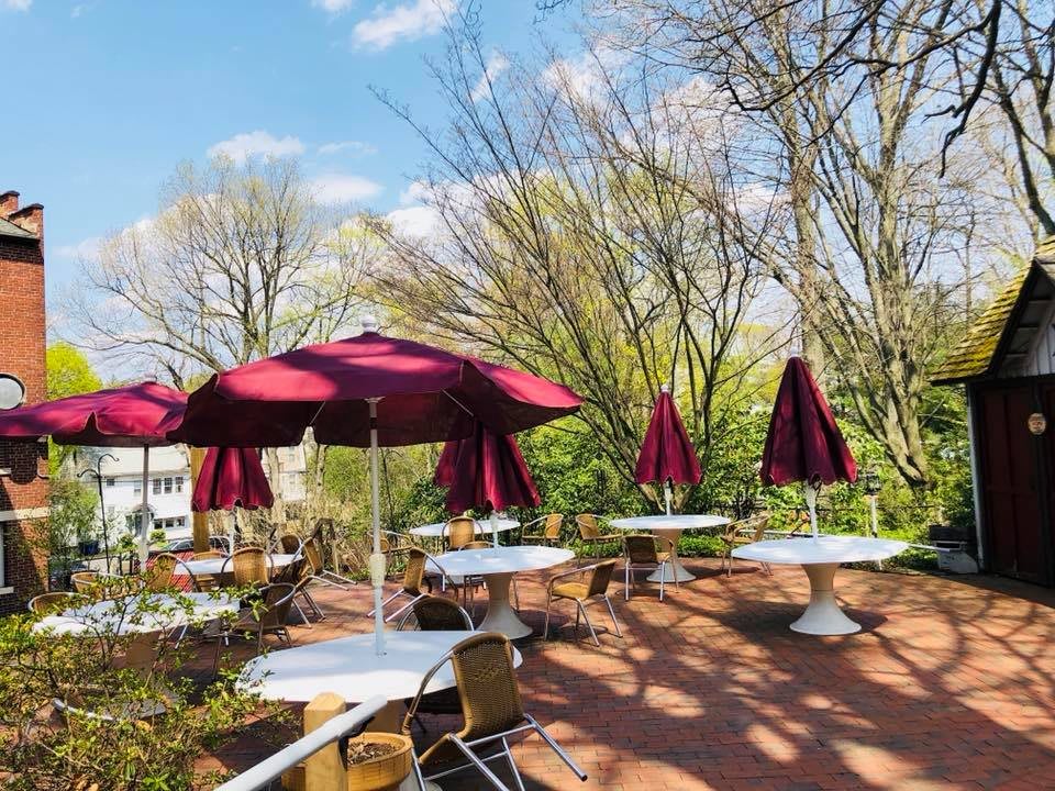A picturesque outdoor patio with tables and chairs under red umbrellas, surrounded by lush trees.