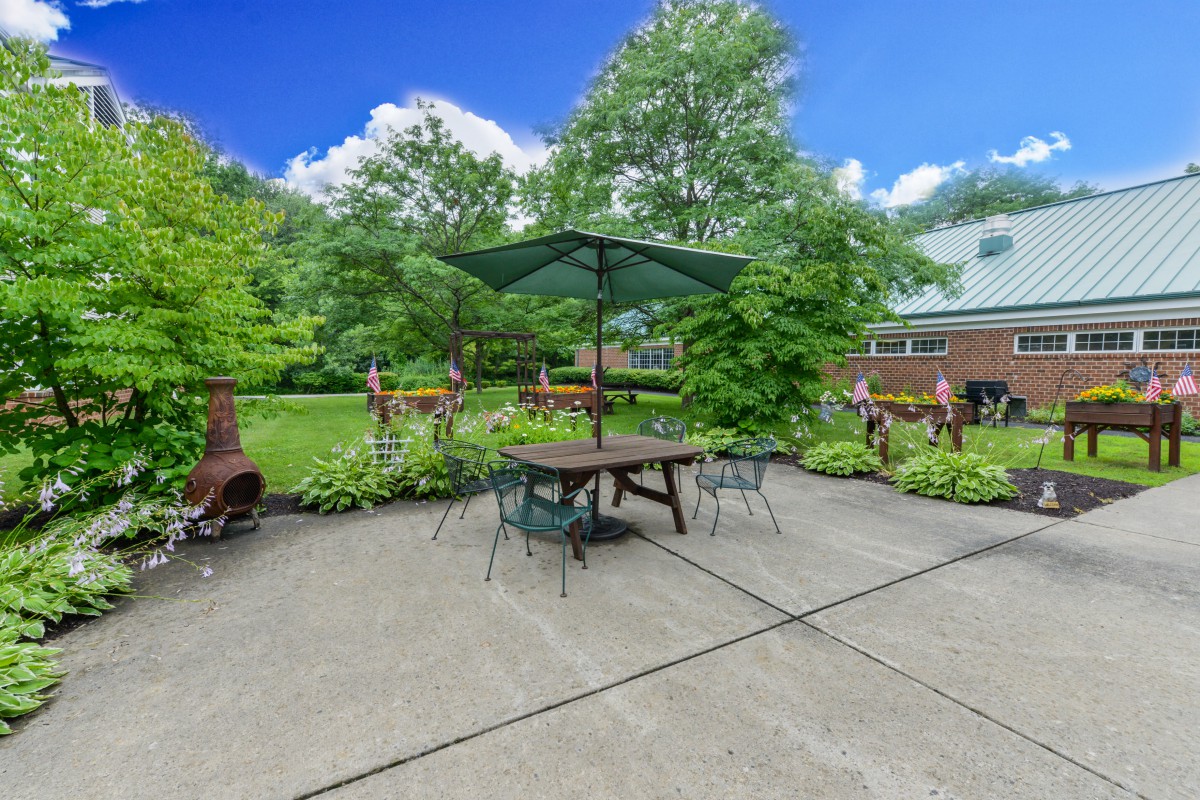 A serene outdoor patio with a table, chairs, and an umbrella, surrounded by a lush garden on a sunny day.