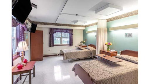 A view of a hospital patient room featuring two beds, a window with curtains, and basic furniture.