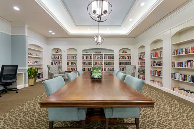 A well-lit library with a large wooden table surrounded by chairs, featuring bookshelves filled with books.