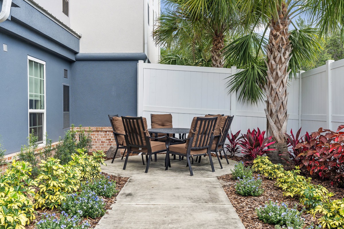 A cozy outdoor patio area with a set of chairs and a table surrounded by lush plants and palm trees.