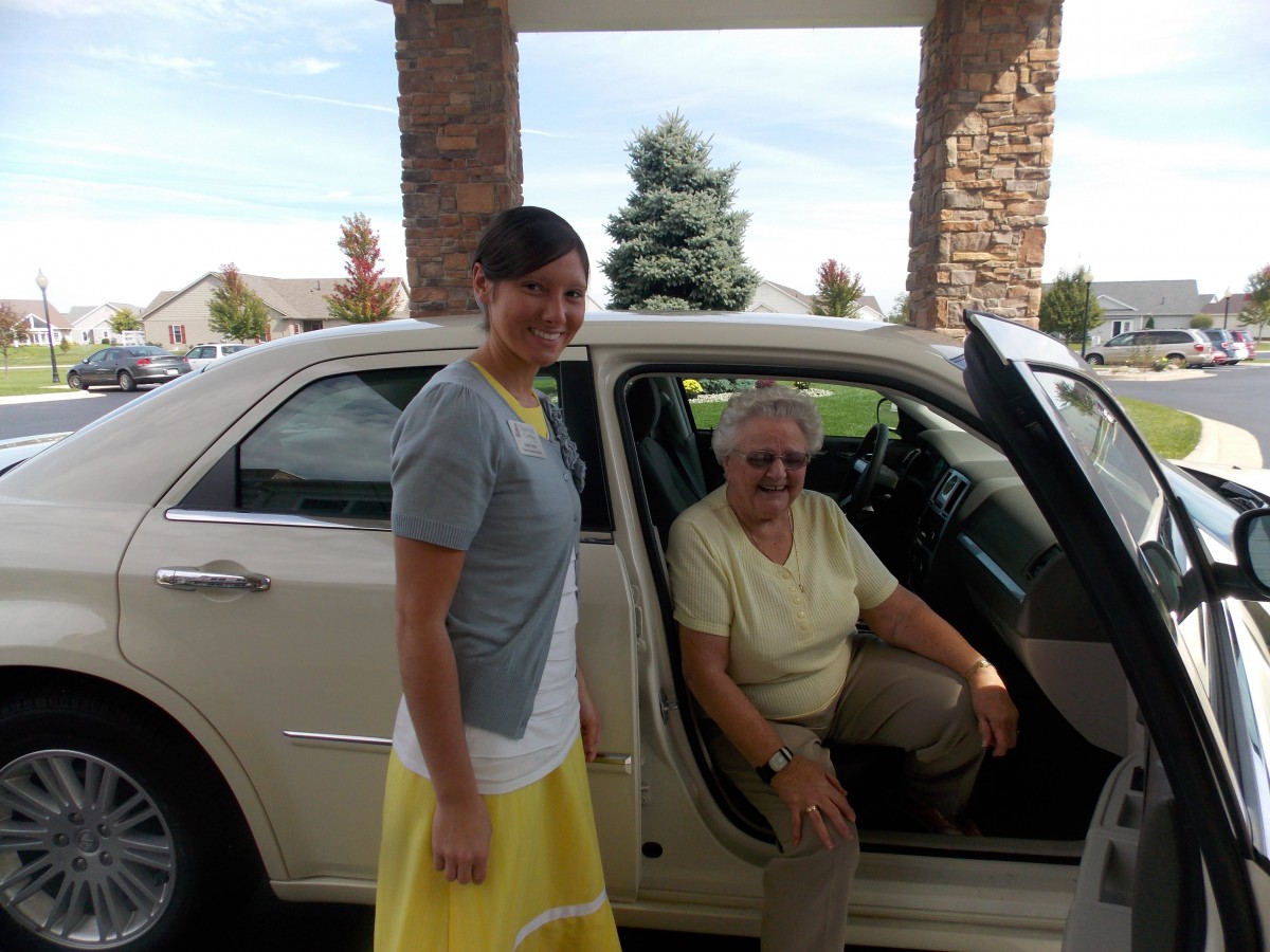 A woman standing next to an elderly person seated in a car.