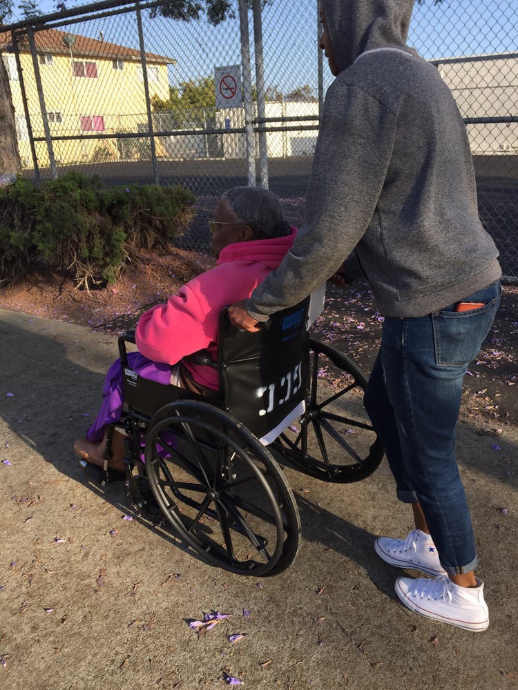 A person assists an elderly individual in a wheelchair outdoors.