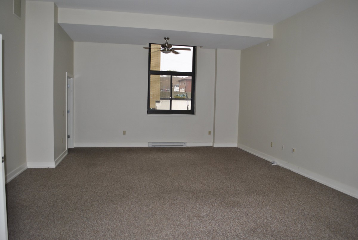 A spacious, empty room with a carpeted floor and a window featuring a ceiling fan.