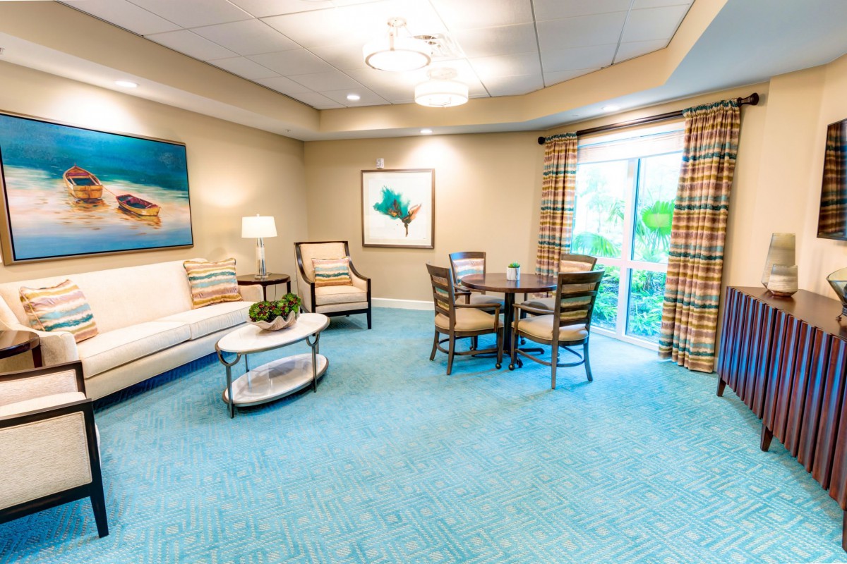 A cozy living room with comfortable seating, a coffee table, and artwork on the walls. Natural light filters through a large window with plaid curtains.