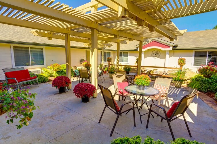 A cozy outdoor patio with a pergola, featuring tables and chairs surrounded by vibrant flower pots under a sunny sky.