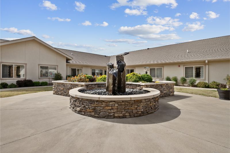 A courtyard with a central fountain surrounded by a stone border, set against a building under a blue sky with clouds.