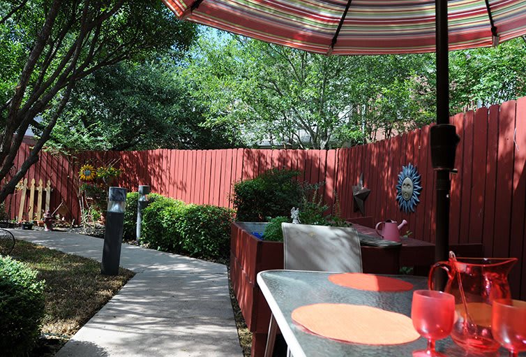 A charming outdoor patio area with a red umbrella, surrounded by lush greenery and a wooden fence.