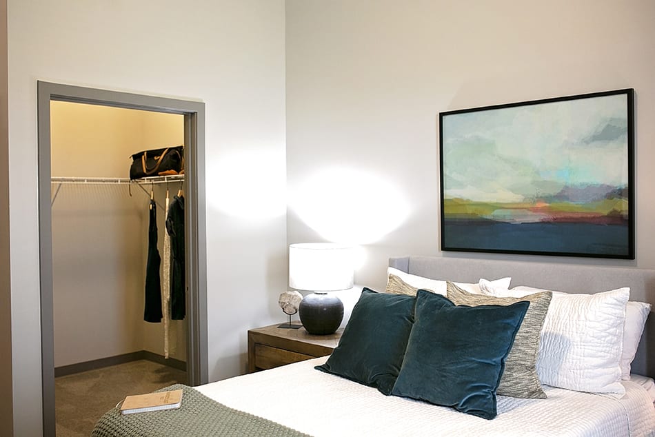 A modern bedroom featuring a neatly made bed with teal pillows, a bedside lamp, and abstract artwork on the wall. An open closet is visible to the side.